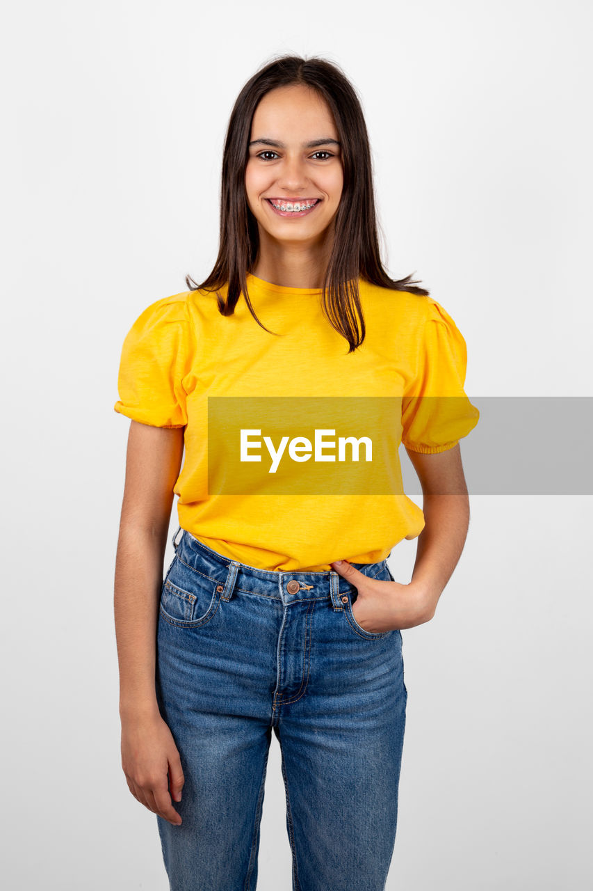smiling, portrait, studio shot, yellow, happiness, one person, looking at camera, casual clothing, women, sleeve, emotion, adult, indoors, t-shirt, young adult, cheerful, long hair, clothing, white background, standing, jeans, pocket, hairstyle, smile, teeth, positive emotion, three quarter length, cut out, front view, relaxation, female, fashion, photo shoot, brown hair, child, copy space, enjoyment, lifestyles, gray, cute, person, joy, hands in pockets, gray background
