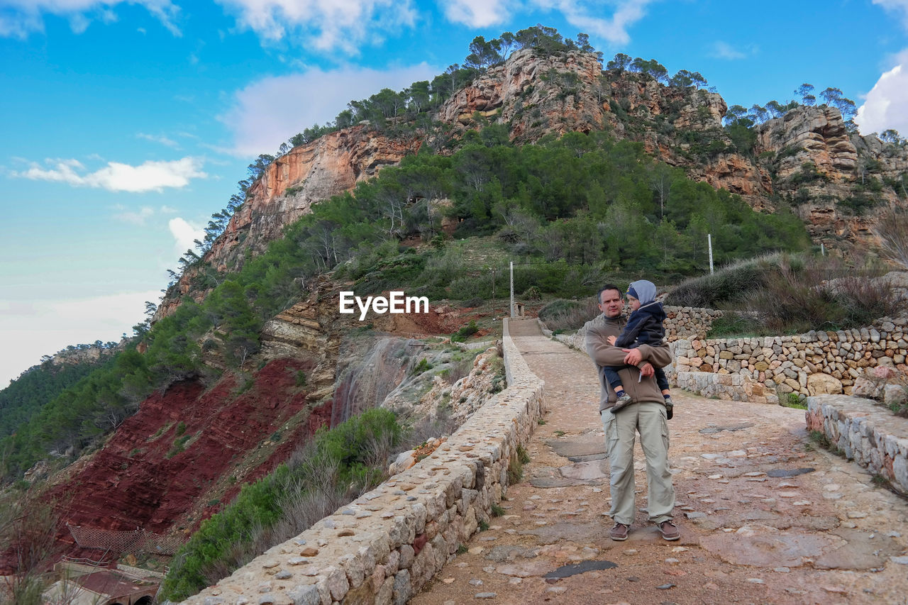 Full length of man carrying son while standing on pathway against mountain