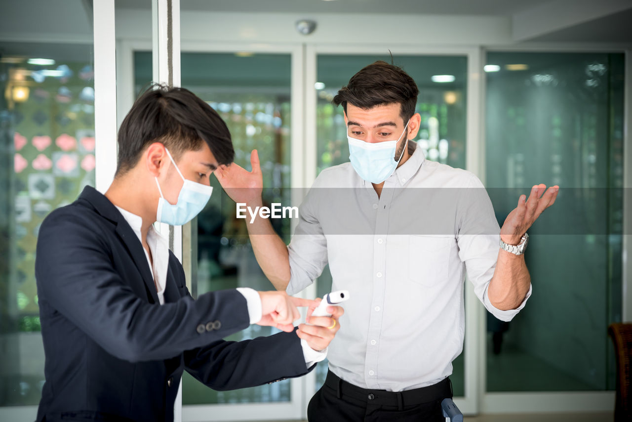 Business person wearing mask showing infrared thermometer to colleague