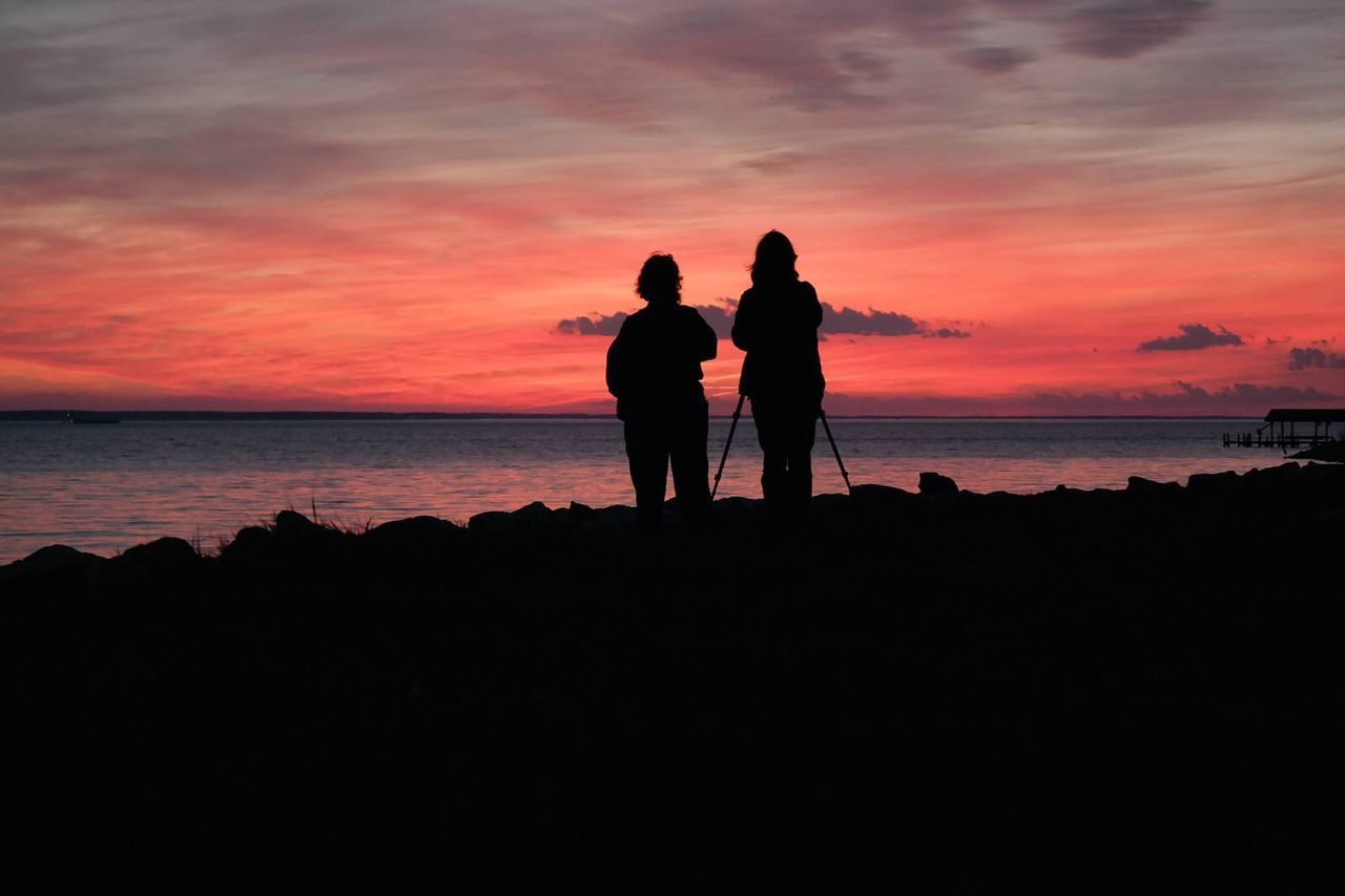 Silhouette friends standing on sea shore against orange sky