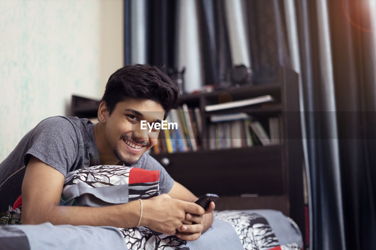 Young indian boy smiling into the camera, while using his phone lying on his bed.