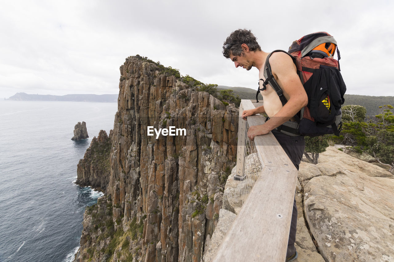 Man carrying a backpack leans over the top of sea cliff in tasmania.