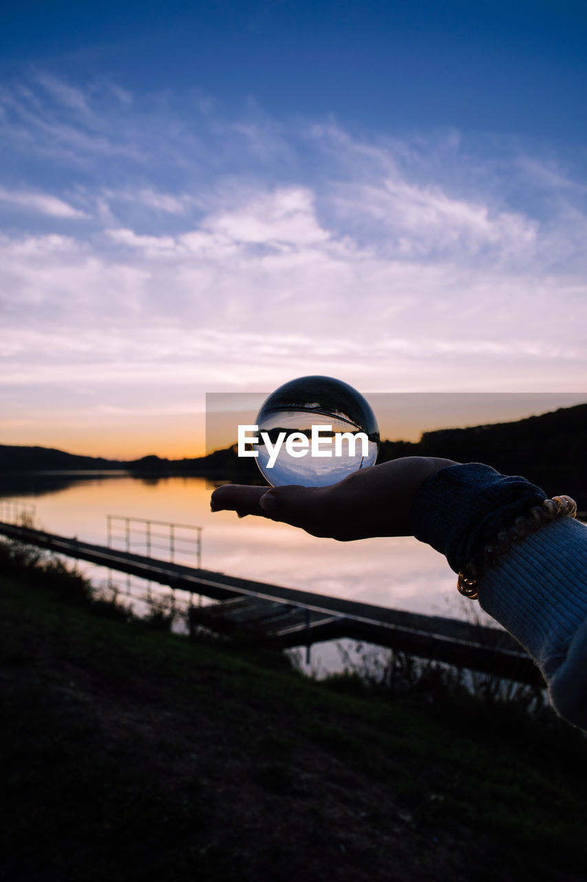 Cropped hand of woman holding crystal ball against sky during sunset