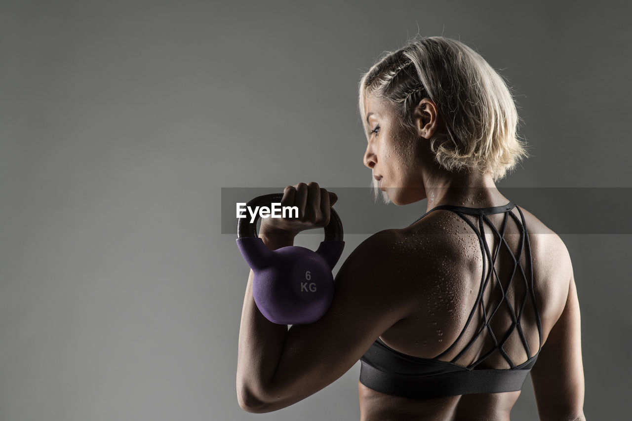 Young woman holding kettlebell while exercising against gray background