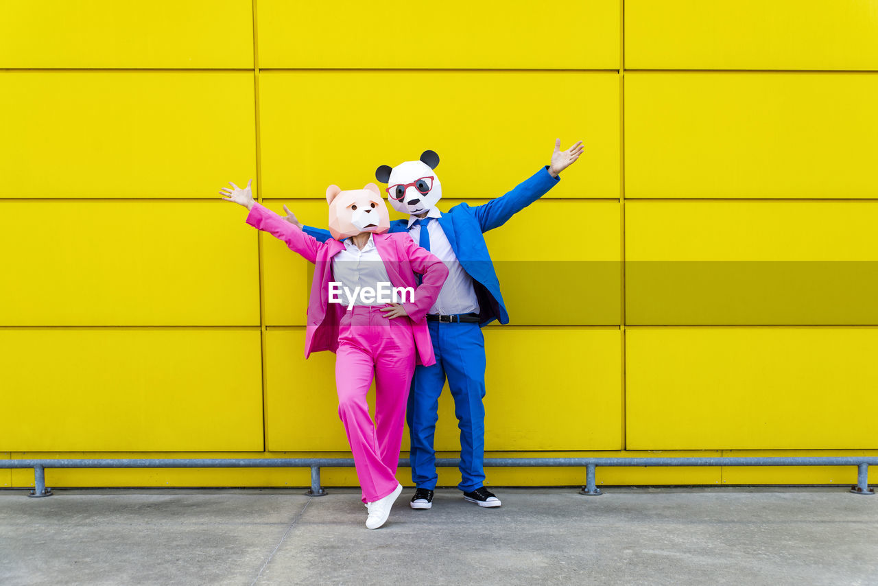 Man and woman wearing vibrant suits and bear masks standing together against yellow wall with raised arms