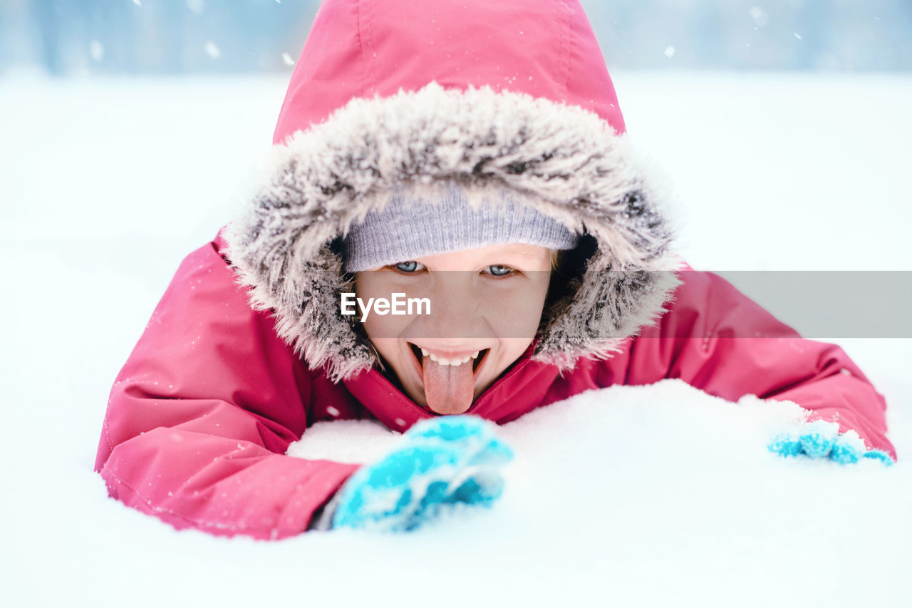 Girl sticking out tongue while lying on snow covered land