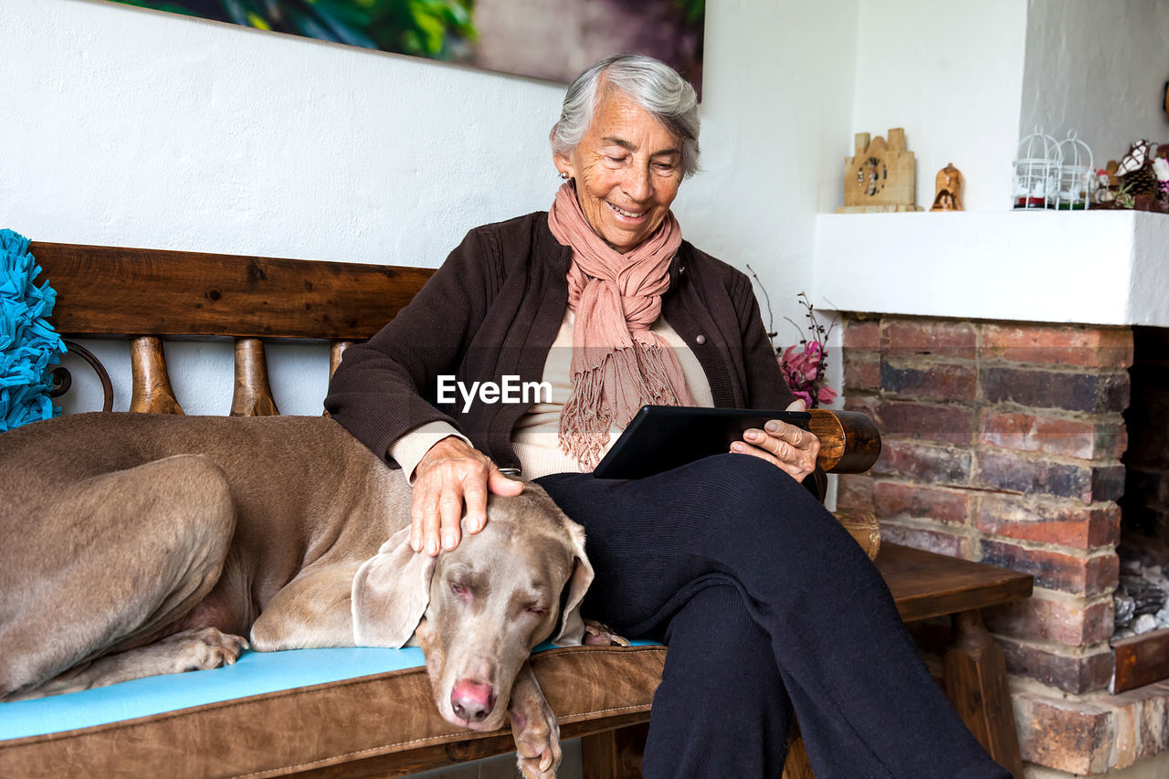 Senior woman using digital tablet with dog sitting on bench at home