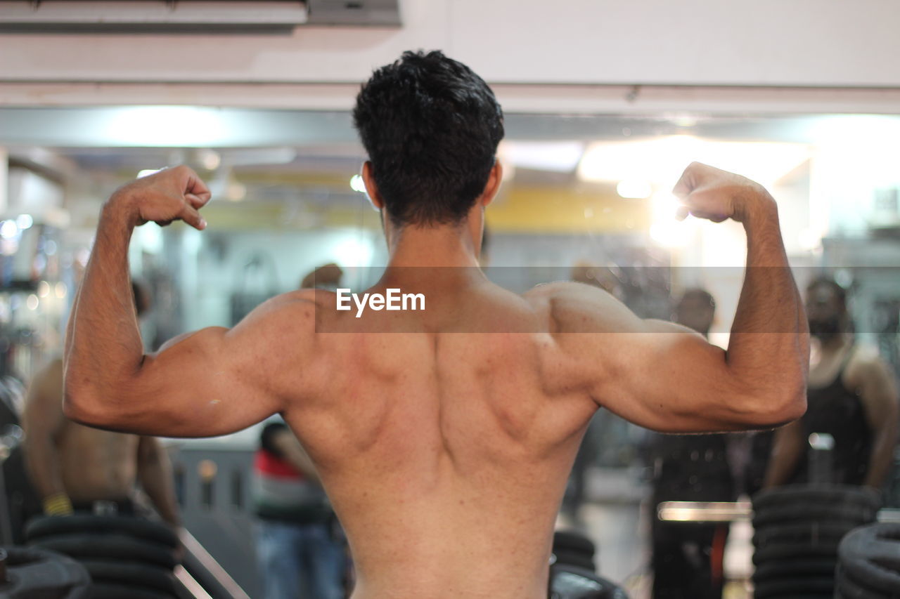 Rear view of shirtless young man flexing muscles at gym