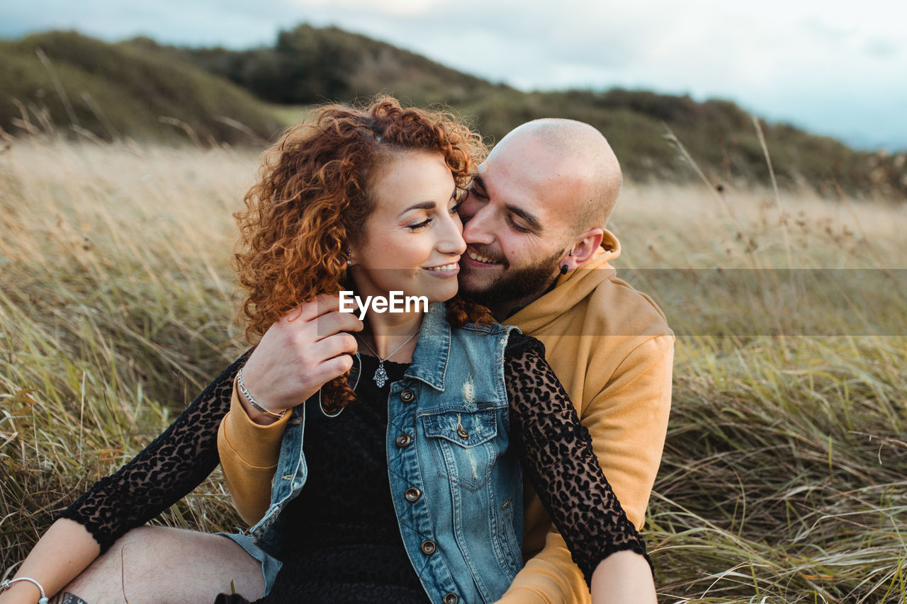 Young bald bearded man in yellow hoodie and jeans shorts embracing happy curly haired girlfriend in dress and denim vest while sitting together on meadow and talking