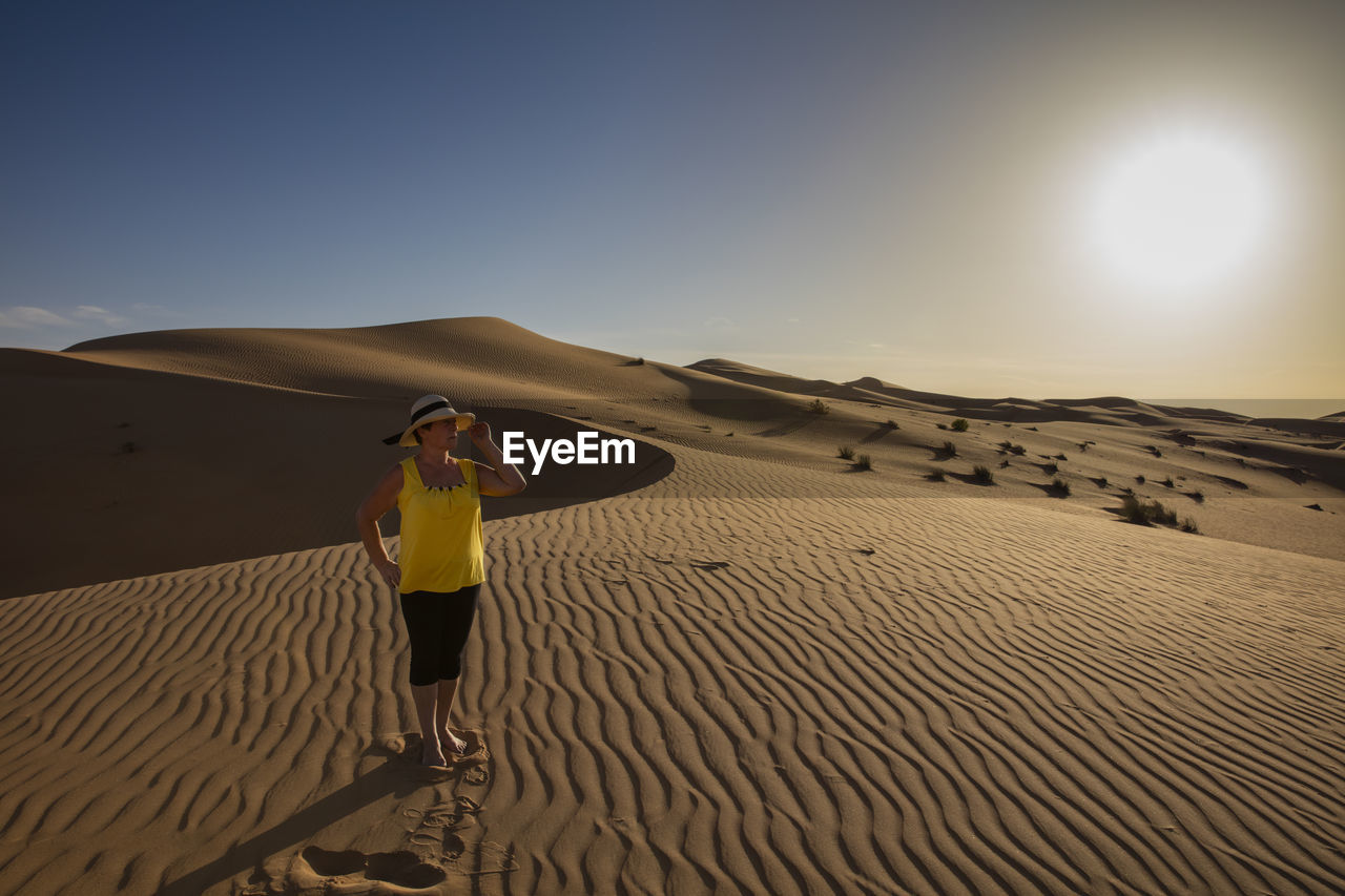 REAR VIEW OF WOMAN STANDING ON SAND DUNE AGAINST SKY