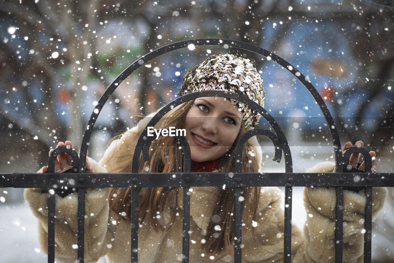 Smiling young woman standing by metal gate during winter