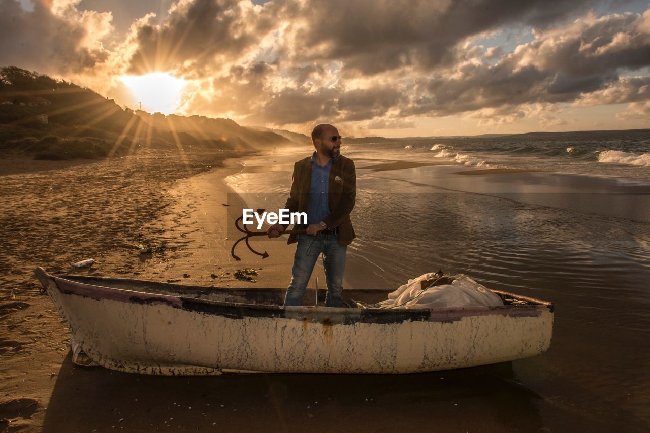 Man holding anchor while standing in abandoned boat at beach during sunset
