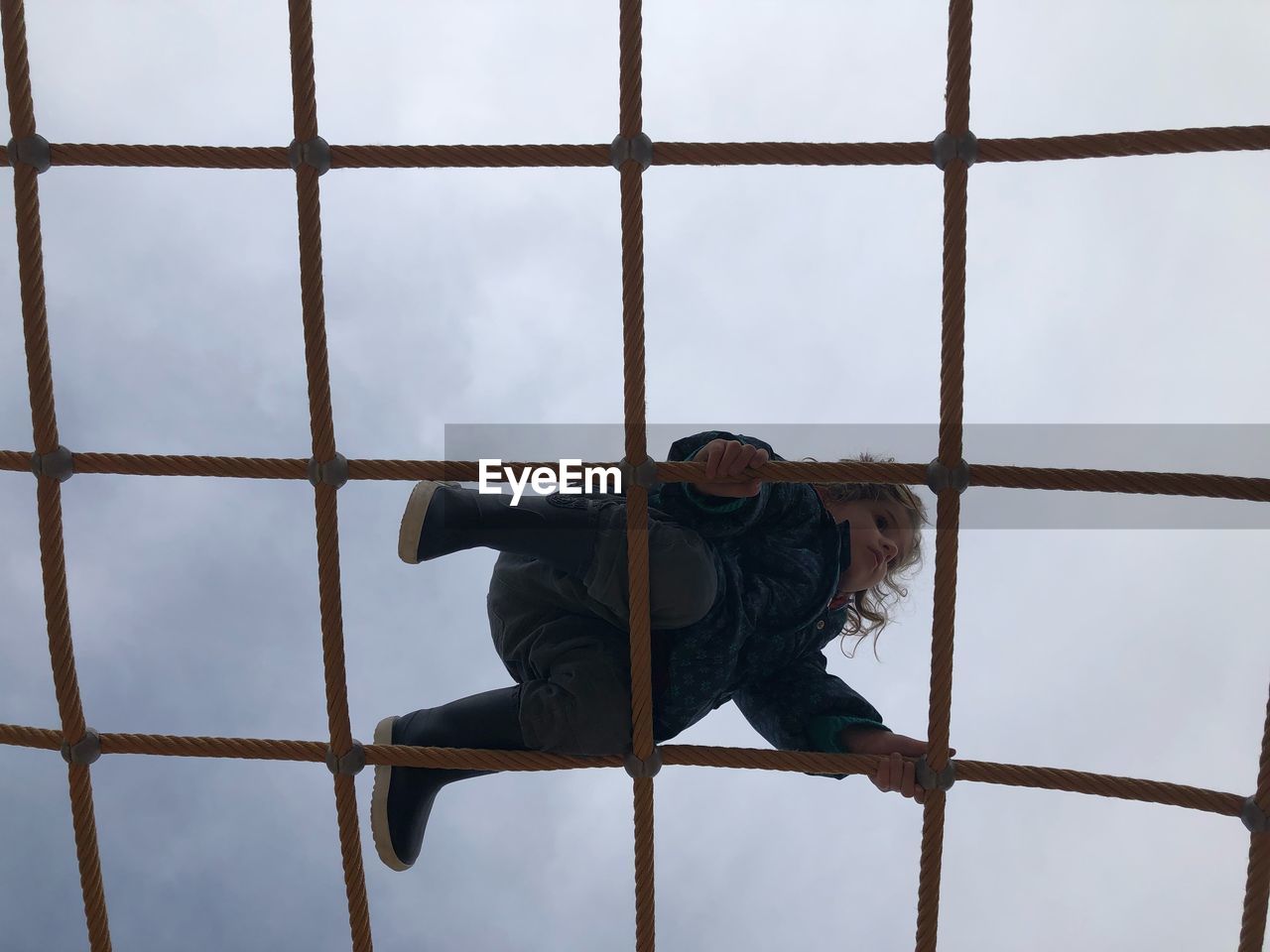 Low angle view of girl climbing jungle gym against cloudy sky during winter