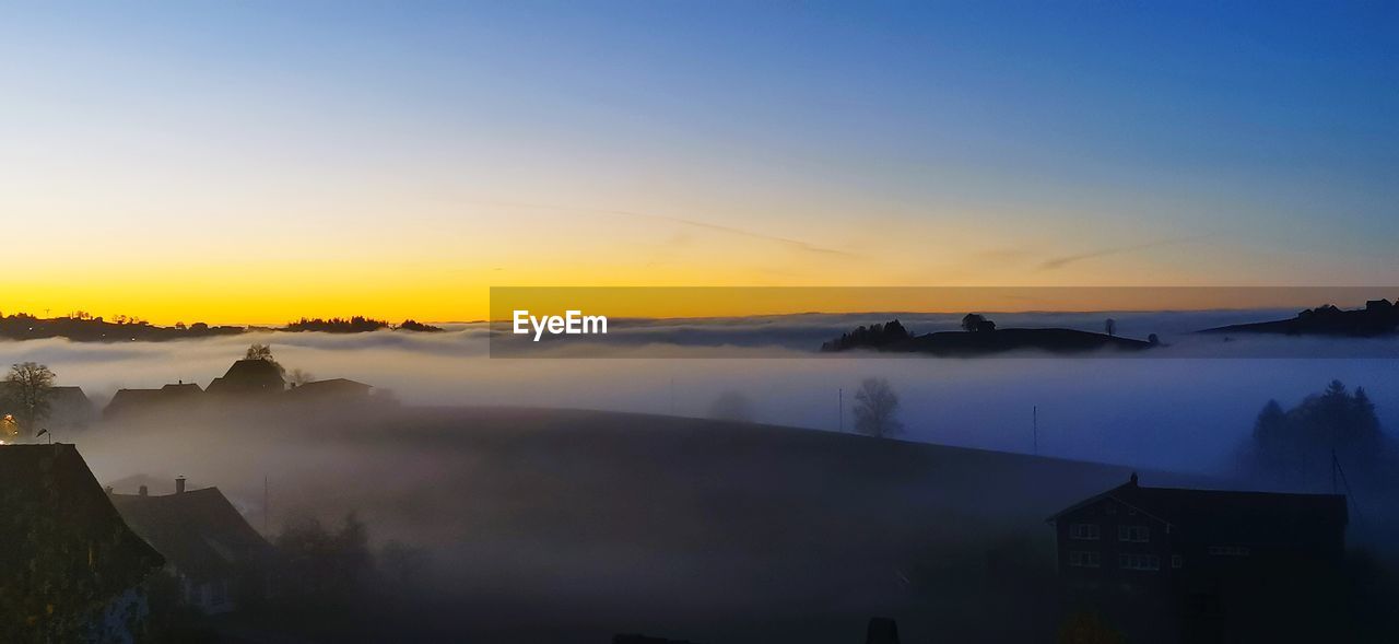 sunrise, dawn, sky, morning, scenics - nature, fog, beauty in nature, environment, water, landscape, nature, tranquility, architecture, sun, mountain, mist, tranquil scene, cloud, twilight, no people, travel destinations, horizon, built structure, idyllic, sunlight, reflection, building, building exterior, land, sea, travel, outdoors, blue, silhouette, house, tourism, tree, non-urban scene, afterglow