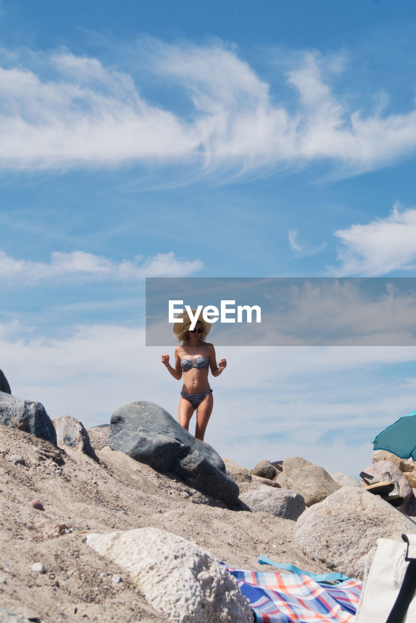 Full length of shirtless woman with bikini on rock at beach against sky