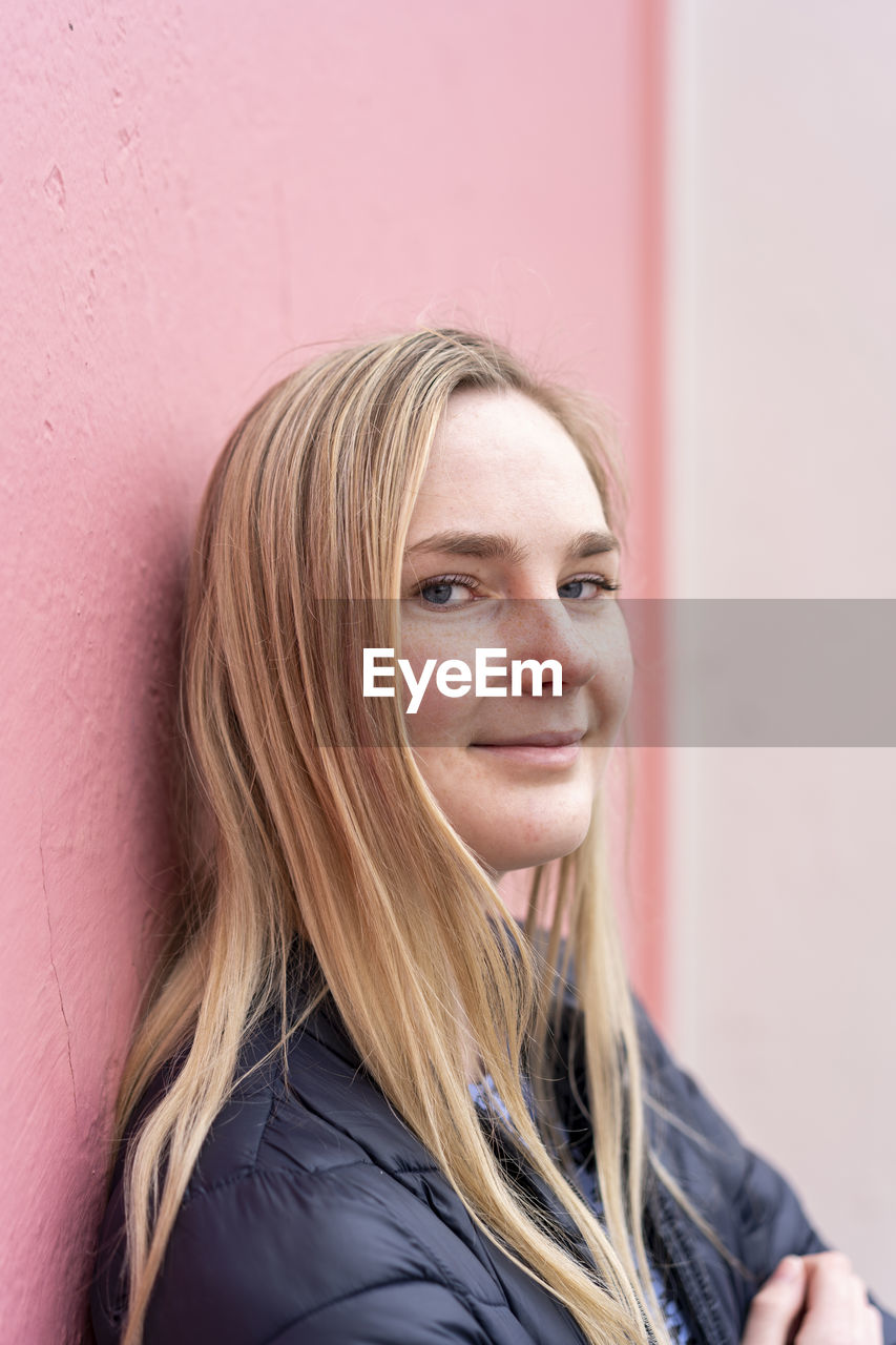 Young smiling woman looking at camera in front of wall outdoors