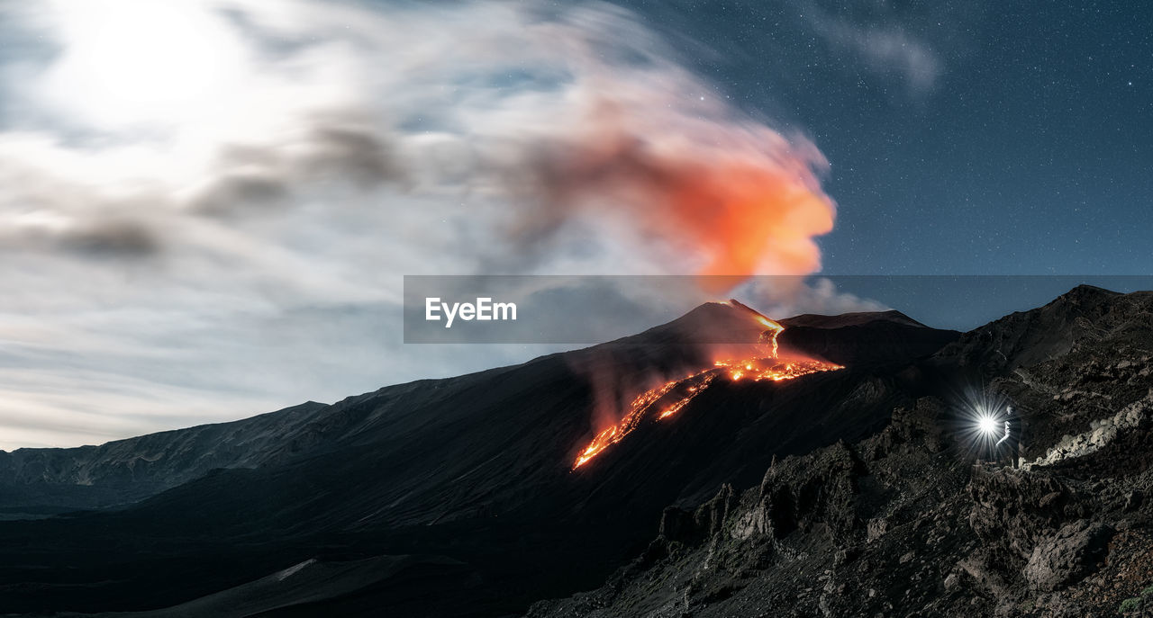 Panoramic image of volcano etna eruption at night with full moon. man with a torch lighting up 