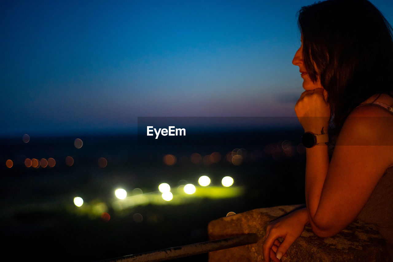 SIDE VIEW OF WOMAN SITTING AT SEA AGAINST SKY AT NIGHT