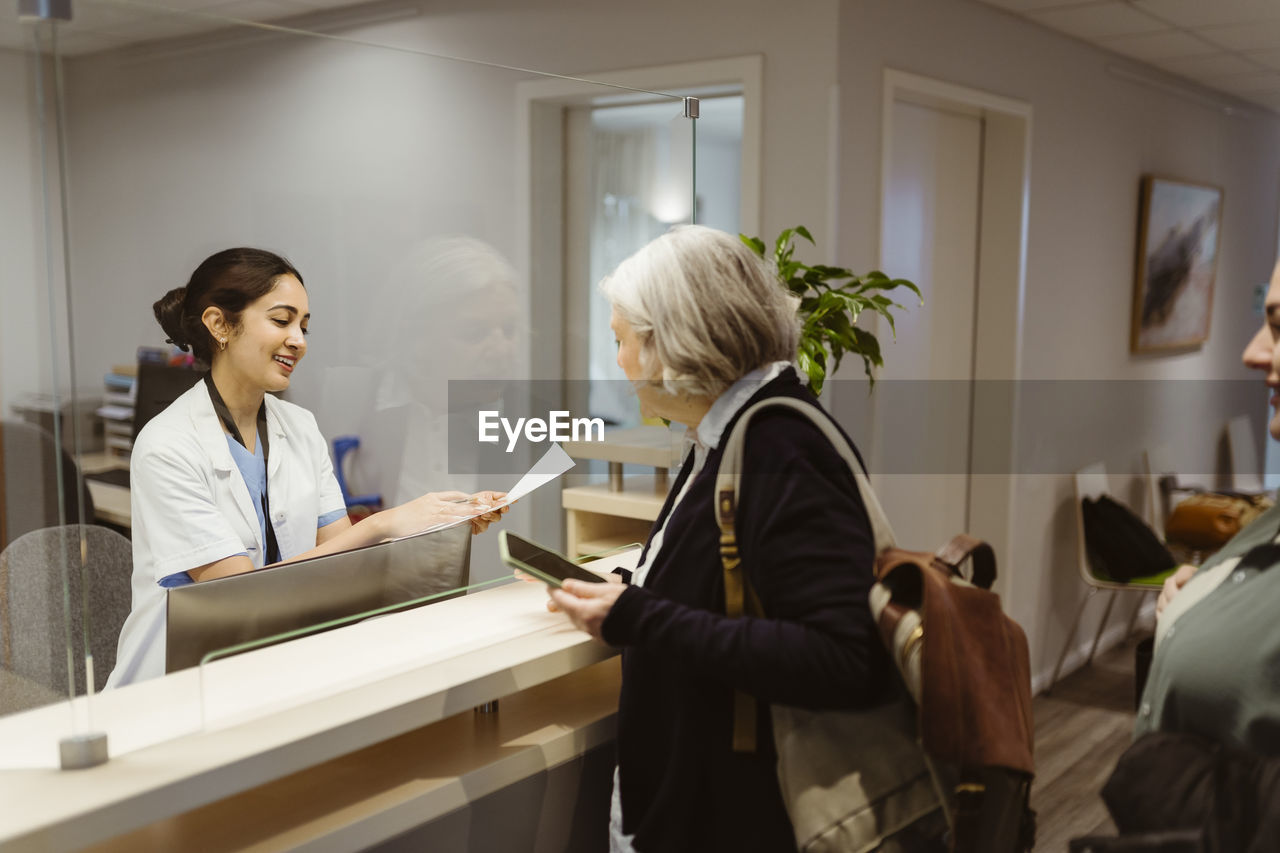 Smiling receptionist explaining form filling procedure to patient through transparent shield in clinic