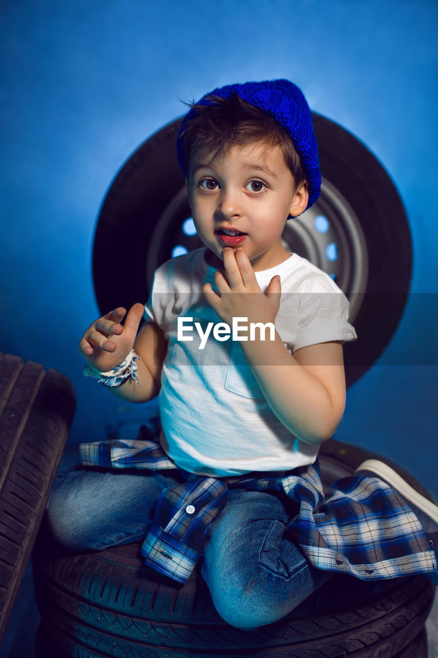 Boy in a white t shirt shirt and hat sits on a background of car wheels on a blue