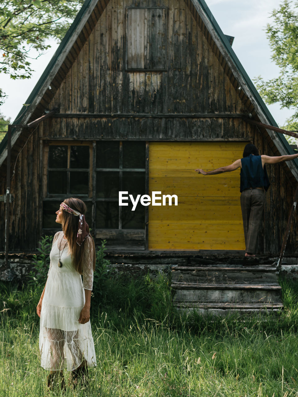 Close-up of a attractive girl looking away and in the background a man balancing on the porch of an abandoned wooden house