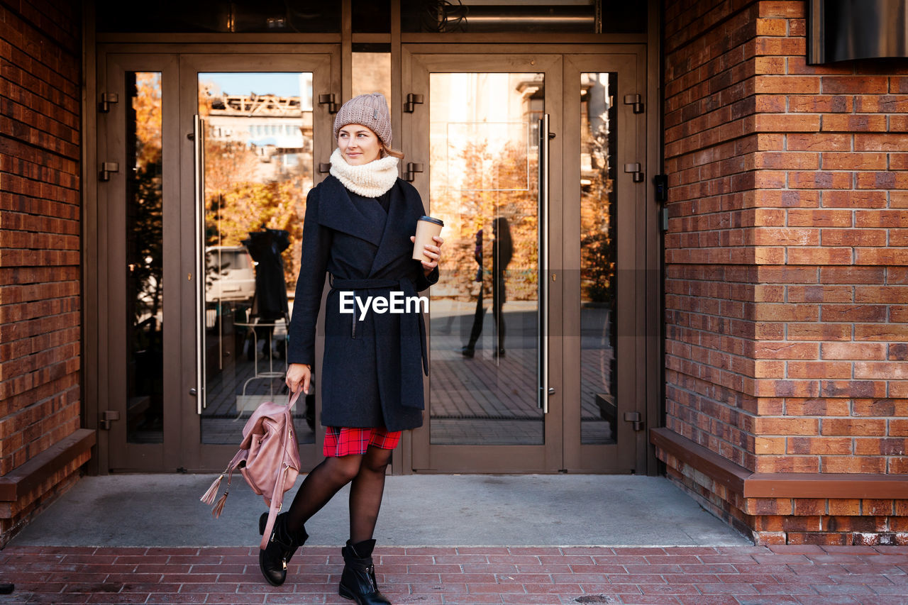 Young female student in warm autumn clothes coming out of the house with a backpack with books 