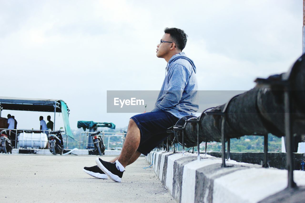 Side view of thoughtful young man sitting on railing against sky