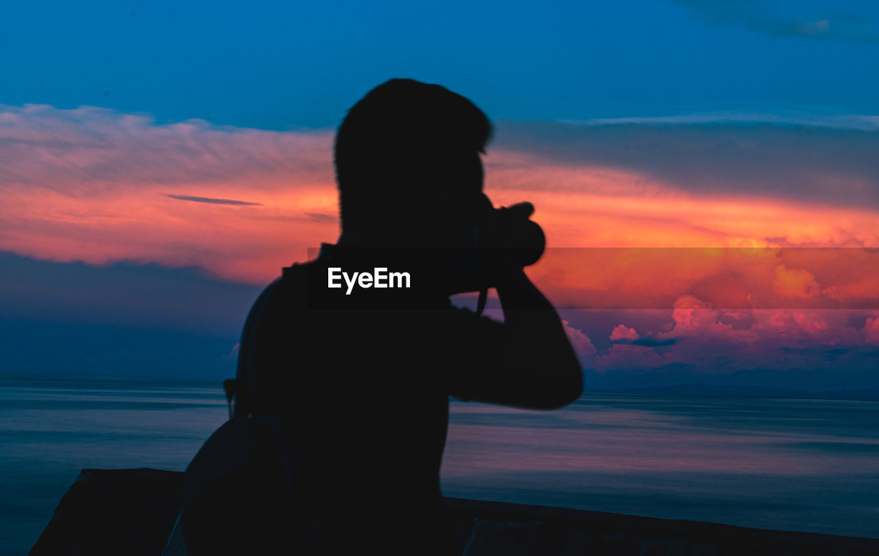 SILHOUETTE MAN PHOTOGRAPHING SEA AGAINST SKY AT SUNSET