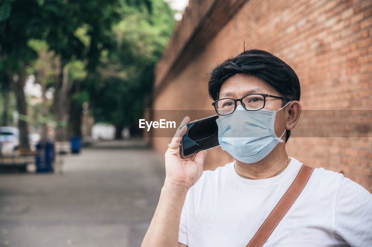 Man wearing mask talking over smart phone while standing against brick wall