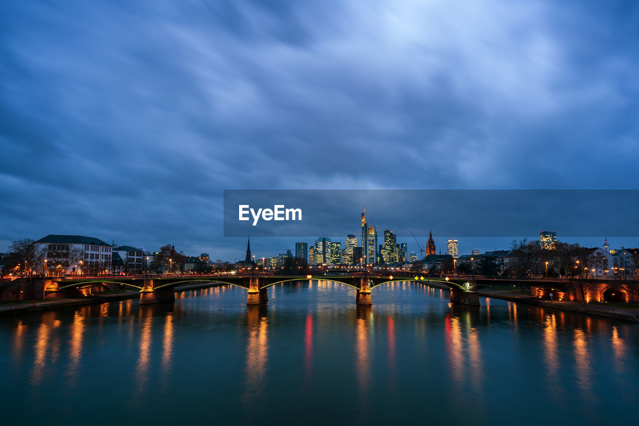 BRIDGE OVER RIVER BY ILLUMINATED BUILDINGS AGAINST SKY AT NIGHT