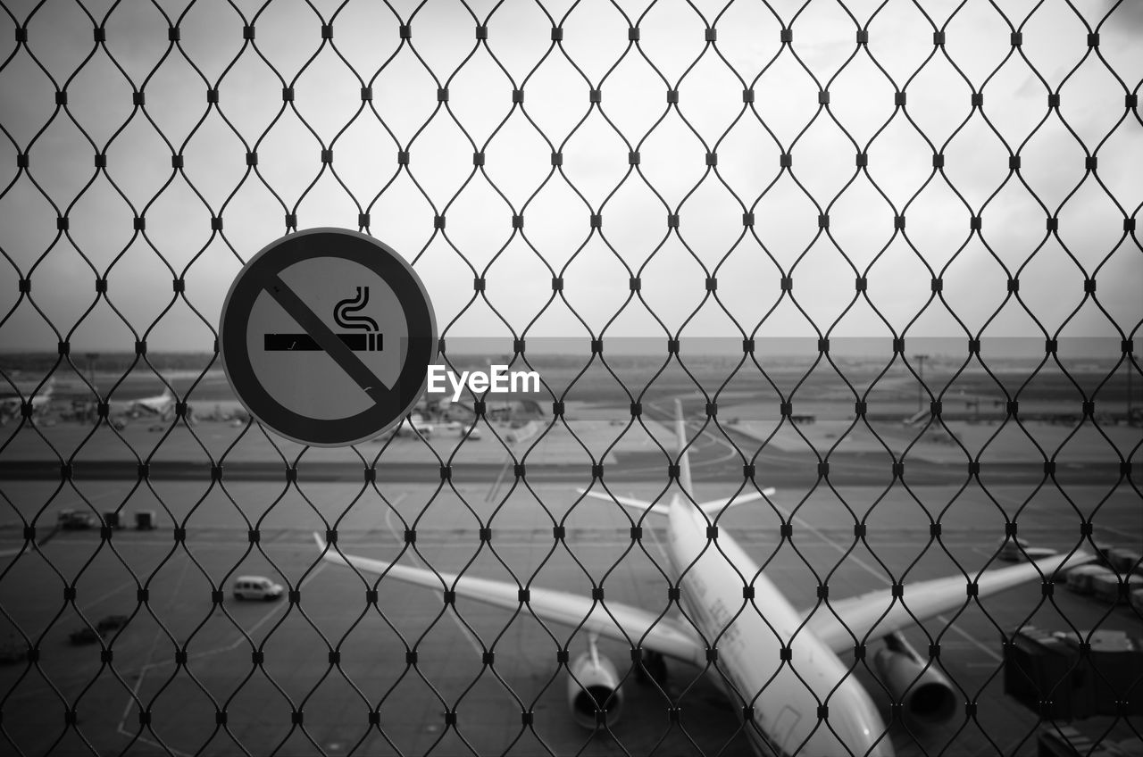 Close-up of signboard on chainlink fence at airport against sky