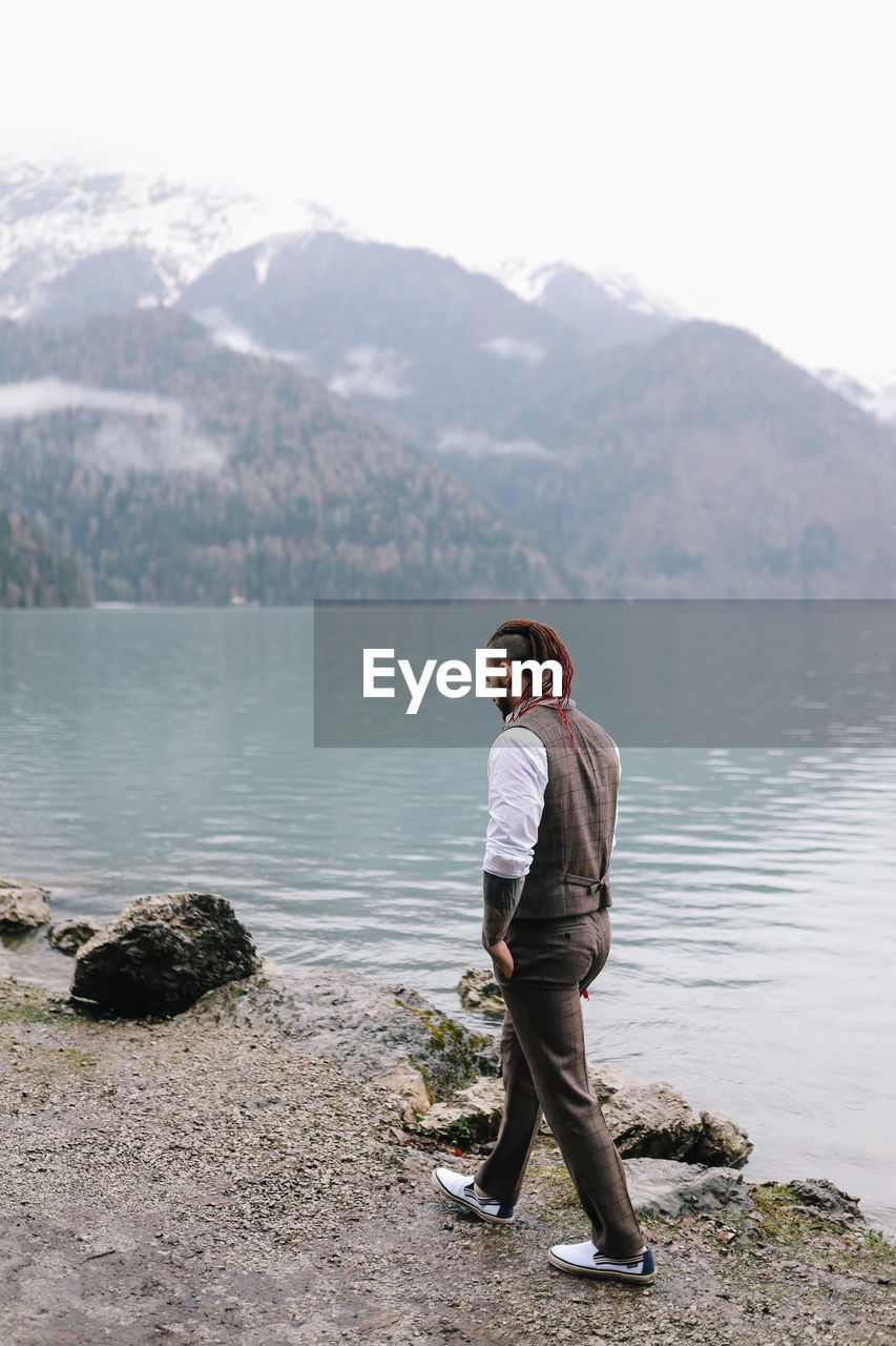 A brutal male hipster groom in a wedding suit in nature by the mountains and lake