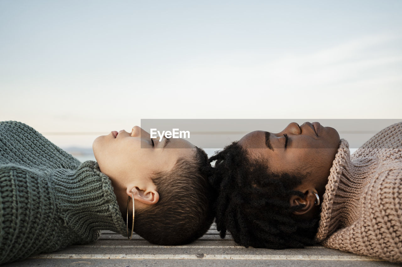 Female friends with eyes closed lying on bench against sky
