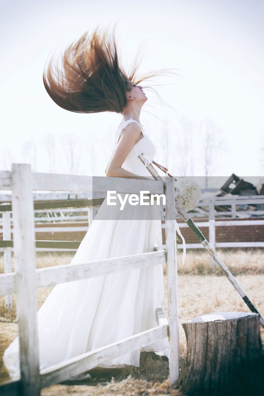 Beautiful bride with tousled hair standing by railing in pen against clear sky