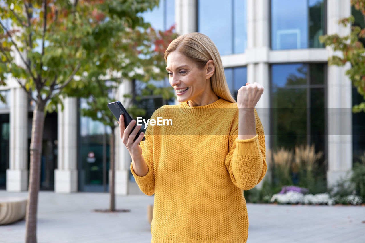 wireless technology, smiling, one person, women, adult, portable information device, smartphone, mobile phone, yellow, technology, happiness, communication, architecture, city, blond hair, telephone, young adult, clothing, emotion, cheerful, lifestyles, spring, standing, city life, holding, casual clothing, street, outerwear, day, smile, outdoors, building exterior, teeth, enjoyment, using phone, built structure, looking, waist up, front view, leisure activity, hairstyle, portability, female, portrait, sweater, person, nature, fashion, building, dress