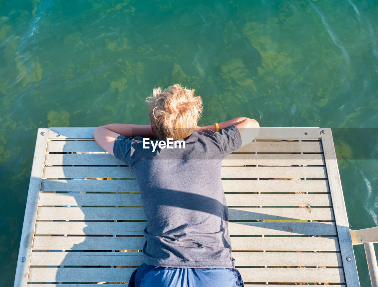 Boy lies on the mole and watches the fish in the lake water below. the joys of childhood