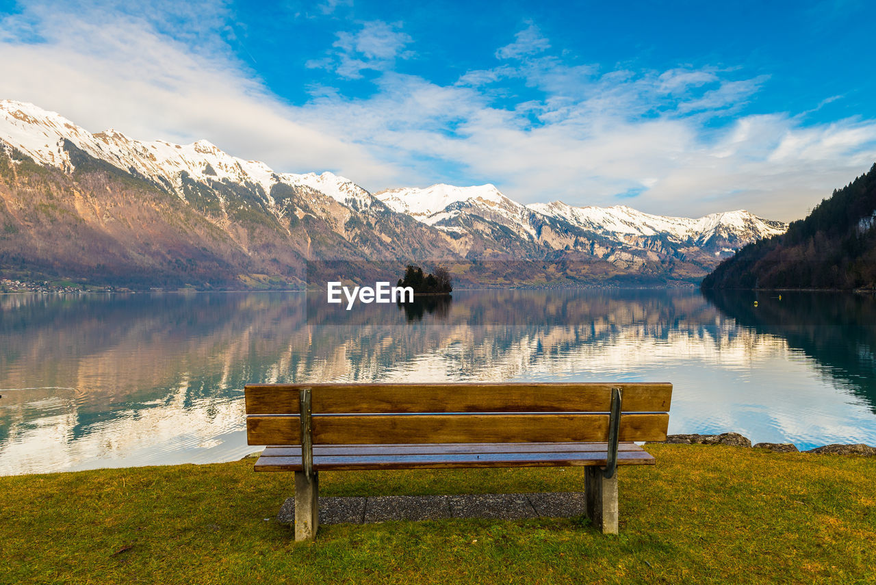 Empty bench at lakeshore against snowcapped mountains