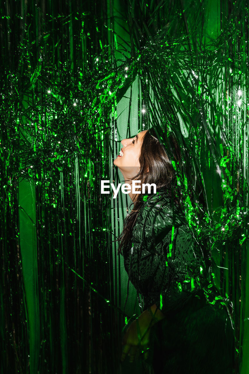 Delighted female wearing festive dress playing with green shiny tinsel stripes at party while looking at camera