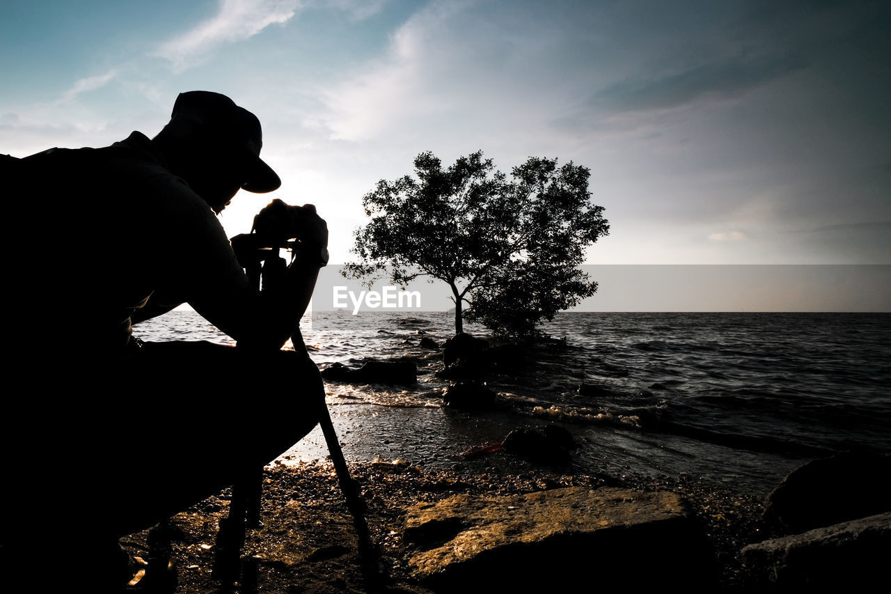 Silhouette person photographing by sea against sky during sunset