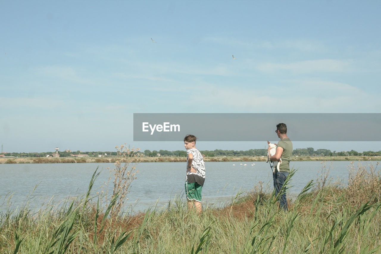 Father and son standing on grassy field by lake against sky