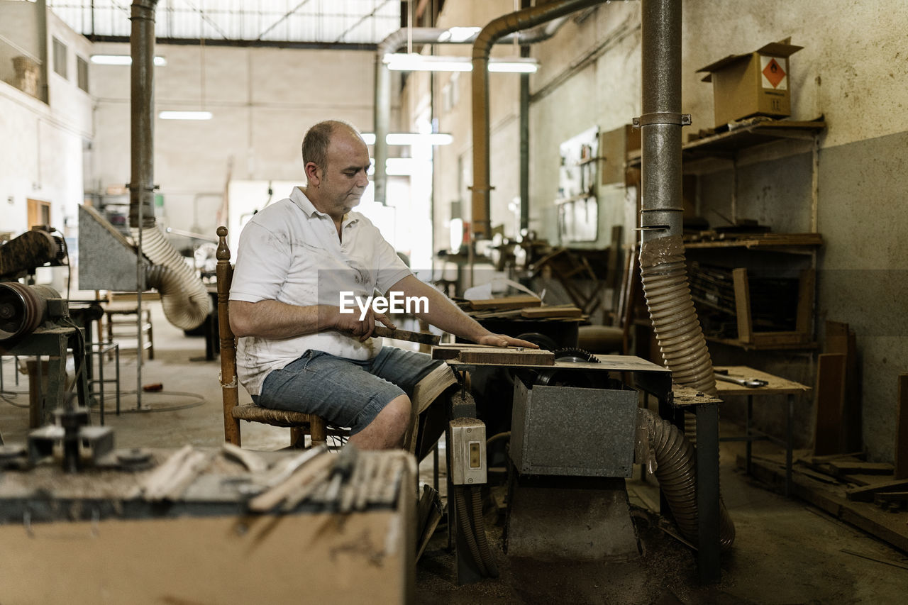 Determined male woodworker sitting at old workbench and working with lumber details in grungy workshop