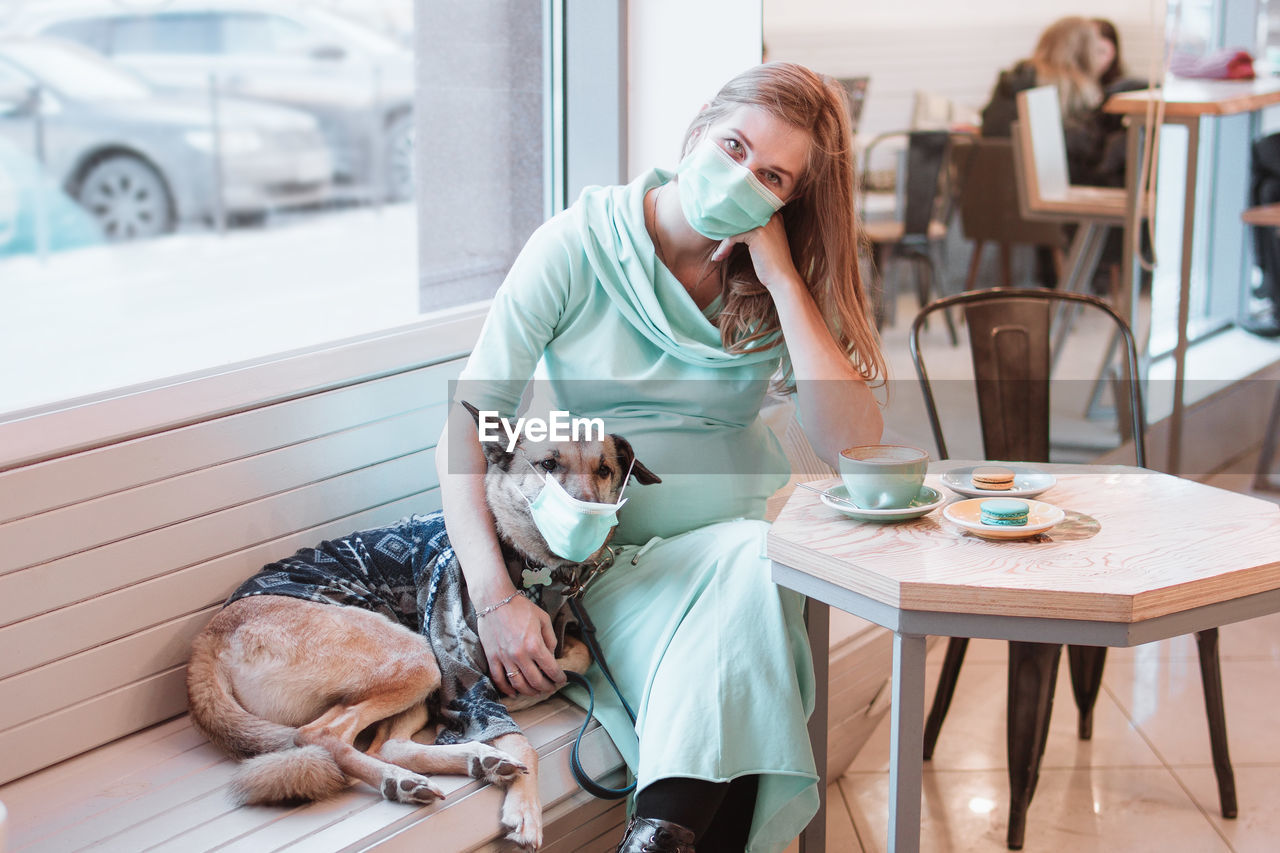 Pregnant women and her dog in protective mask are sitting at the table in cafe. 