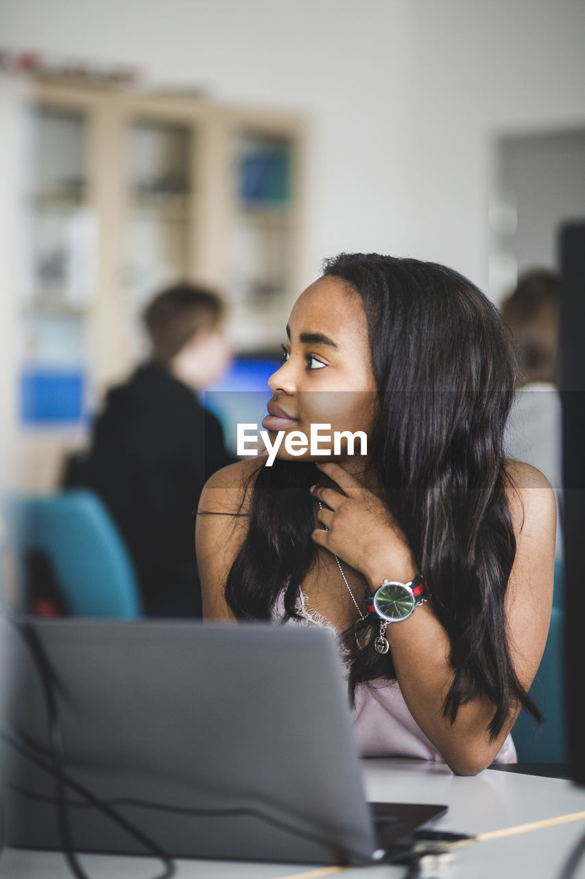 Confident female high school student looking away while sitting with laptop at desk in classroom