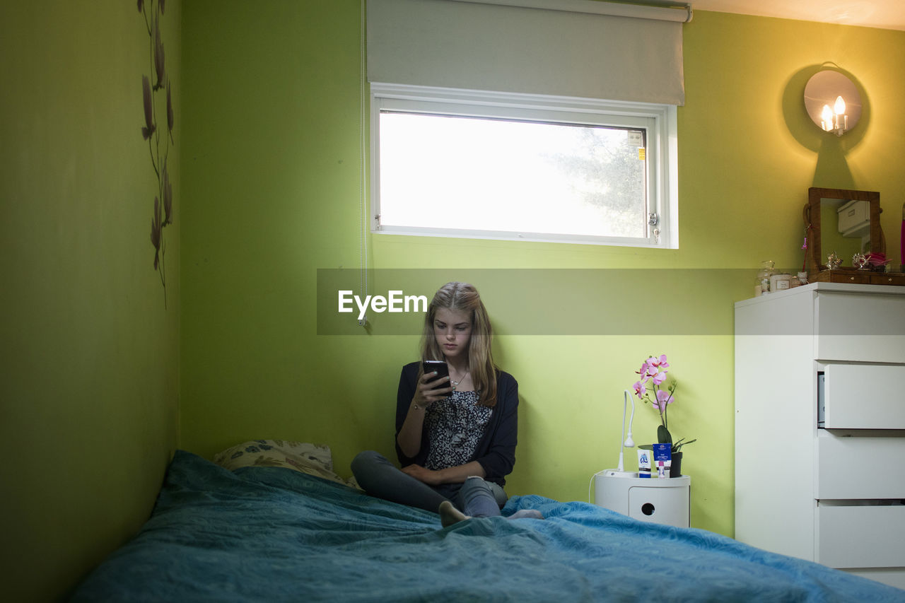 Teenage girl using smart phone while sitting on bed at home