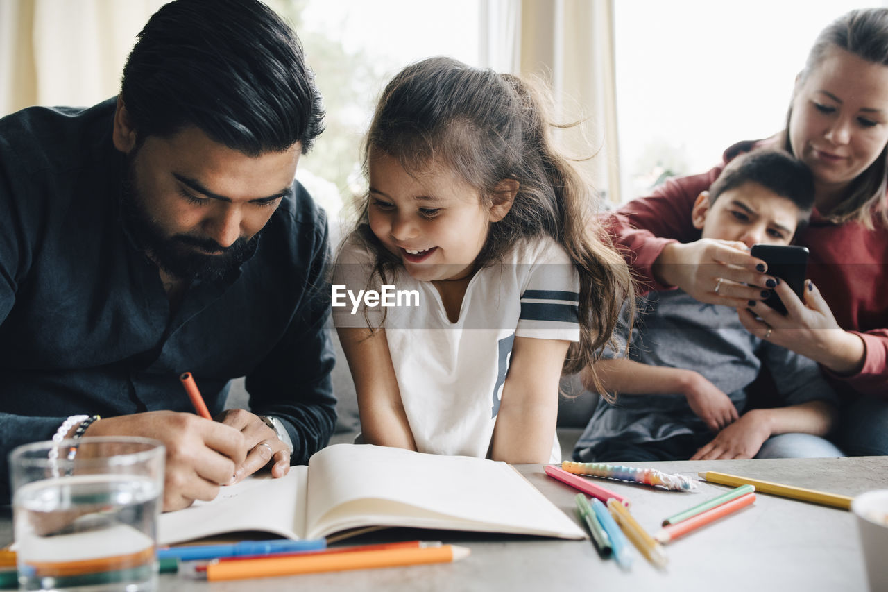 Father teaching drawing to daughter while woman using smart phone with autistic son in background