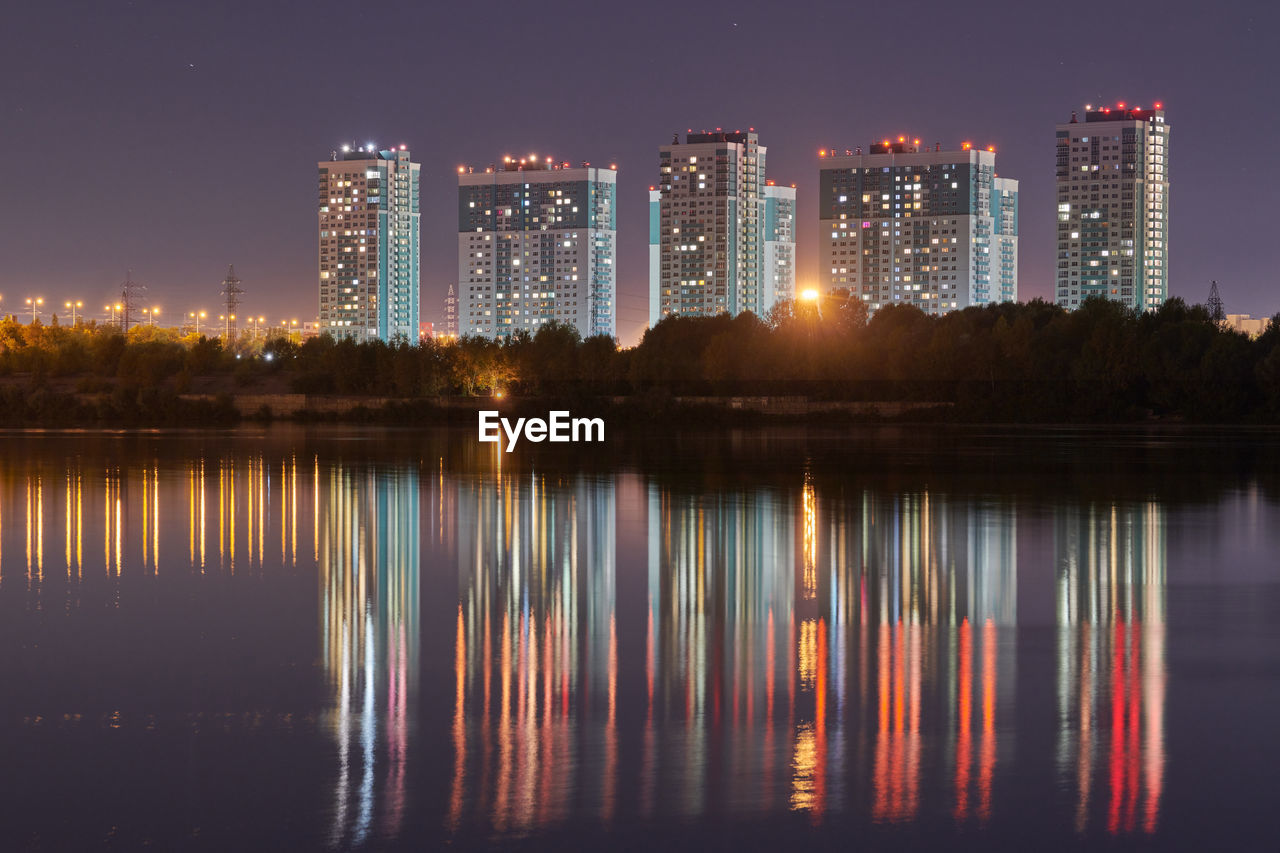 Illuminated buildings by lake against sky in city at night