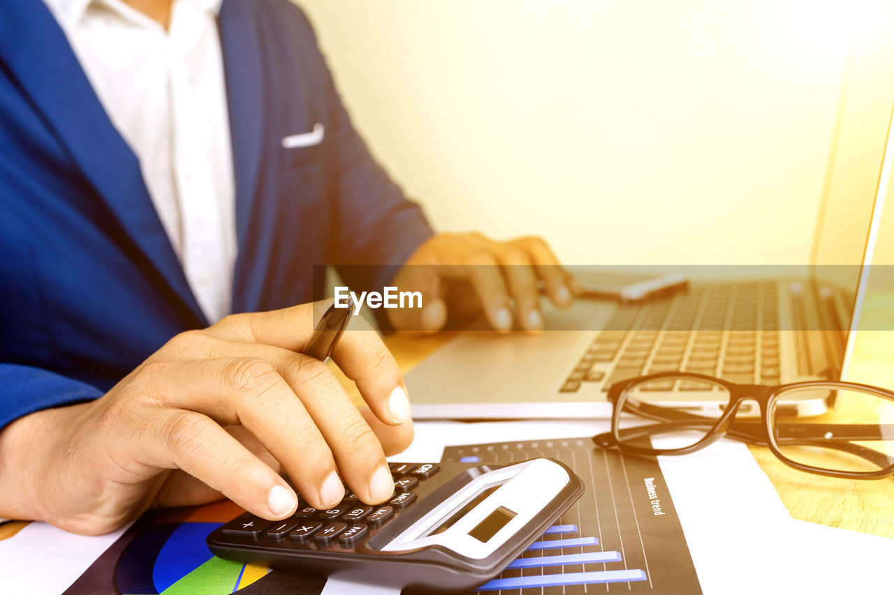 Midsection of man using calculator while typing in laptop on table in office