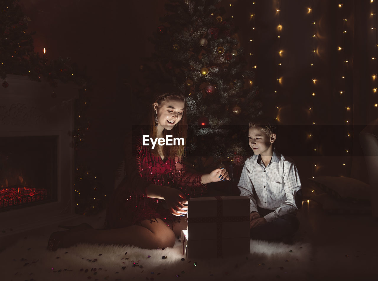 TWO PEOPLE SITTING AT ILLUMINATED CHRISTMAS TREE AT NIGHT