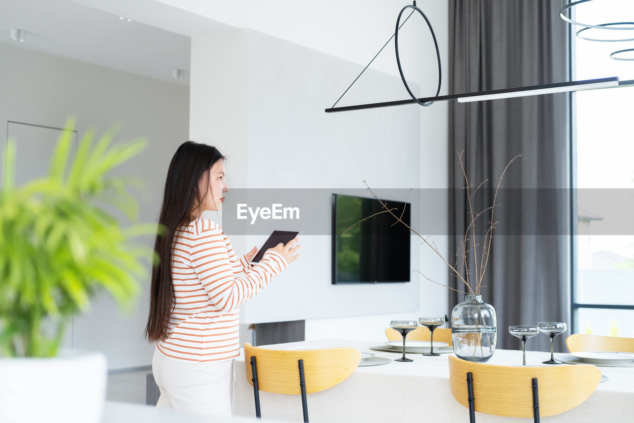 Asian woman controlling smart home functions through tablet at modern home, looking at ceiling lamp