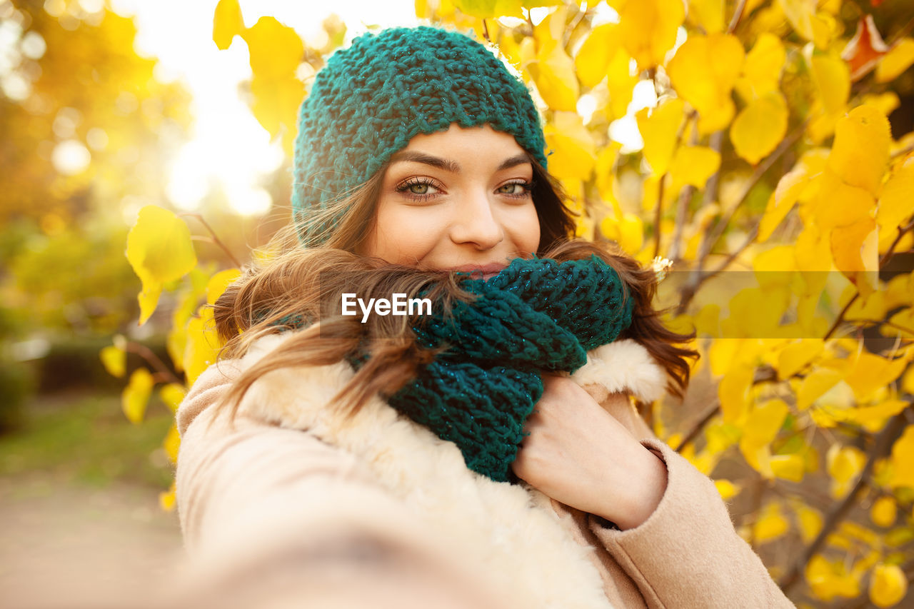 Portrait of beautiful young woman in warm clothing against autumn tree